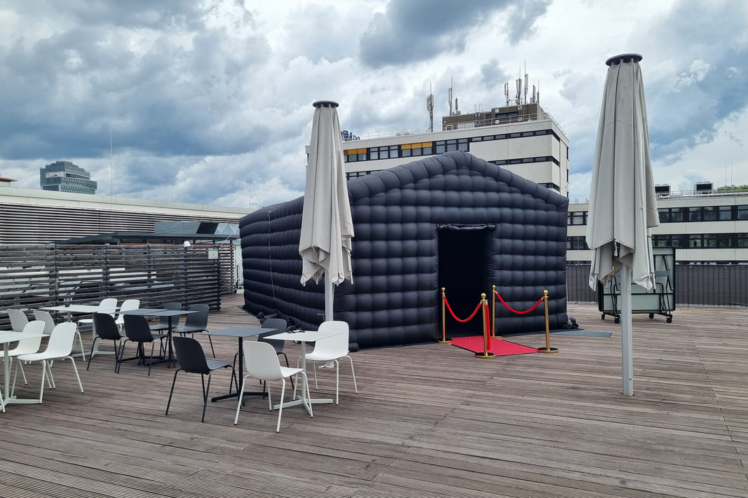 Black inflatable structure with red carpet and gold stanchions on a rooftop terrace with tables, chairs, and closed umbrellas under a cloudy sky.
