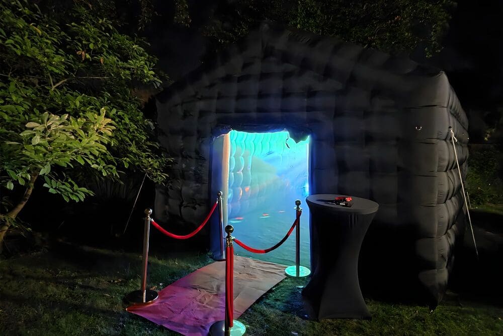 Black inflatable structure at night with glowing blue entrance, red velvet ropes, and a covered standing table nearby.