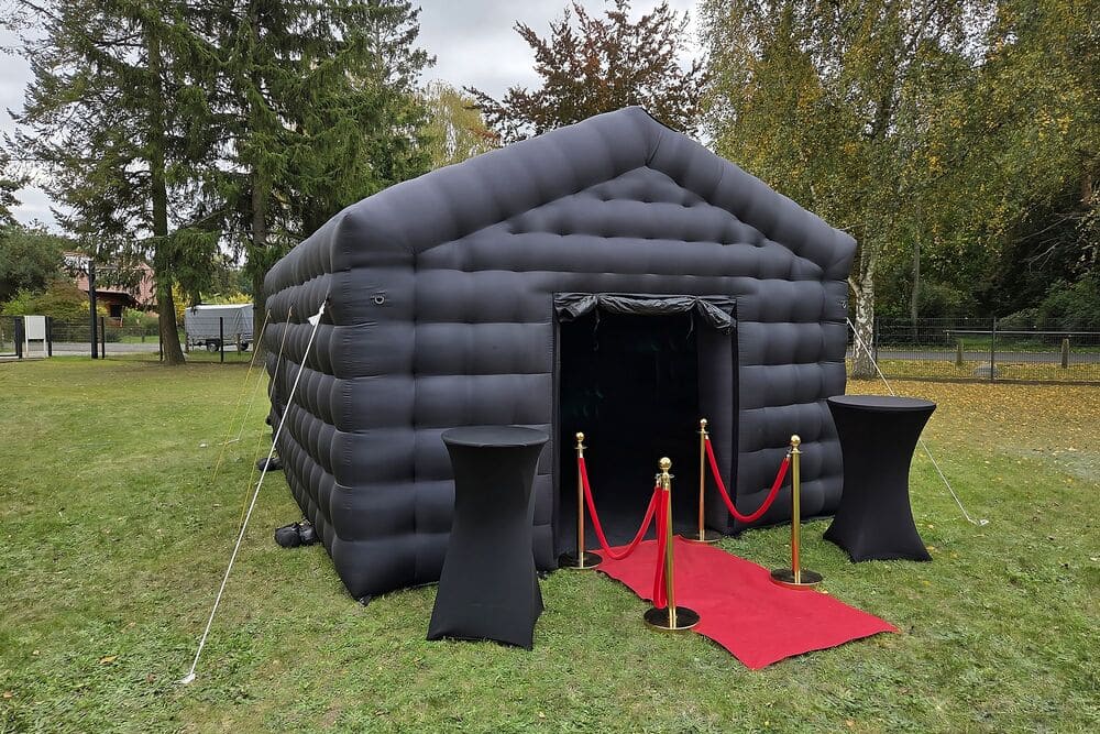 Black inflatable tent with an entrance featuring red carpet and gold stanchions with red ropes on a grassy outdoor area.